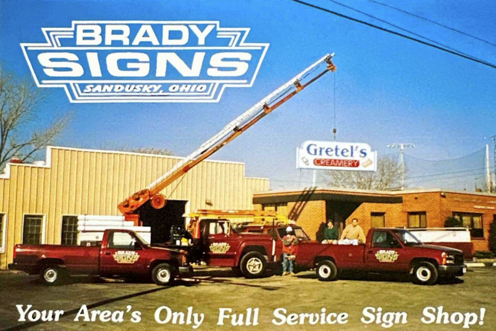 Vintage Brady Signs promotional photo with red service trucks and a boom crane outside the Sandusky, Ohio sign shop